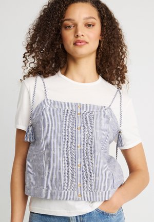 Jeune femme aux cheveux bouclés portant un t-shirt blanc sous un haut sans manches bleu et blanc à rayures avec des boutons et des bretelles à franges.