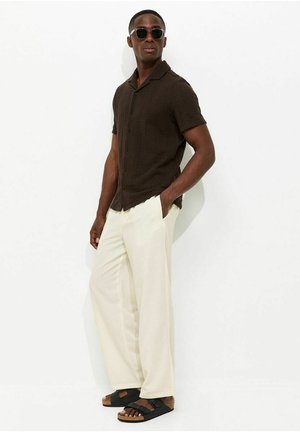 Mężczyzna w ciemnobrązowej koszuli z krótkim rękawem, kremowych szerokich spodniach, czarnych sandałach i okularach przeciwsłonecznych, stojący z jedną ręką w kieszeni.