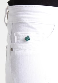 Jean en denim blanc avec un petit élément décoratif turquoise sur la poche arrière, équipé de garnitures argentées et d'une texture lisse.