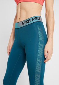 I leggings Nike Pro in teal presentano un'alta vita con una banda riflettente con logo e dettagli decorativi in rete lungo il lato. Tessuto morbido e elasticizzato.