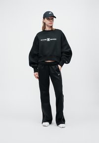 Černá zkrácená mikina s logem "NIKE SHOX", spárovaná s černými tepláky a odpovídajícím kloboukem. Měkká textura, volný střih, nenápadné bílé akcenty.
