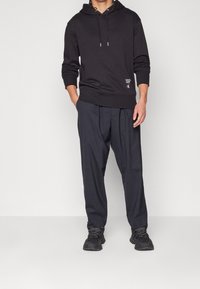 Muž v černé mikině s logem Calvin Klein, tmavých volných kalhotách a černých teniskách stojící s rukama v kapsách.