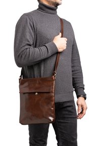 Borsa a tracolla in pelle marrone con una superficie liscia, tasca frontale piatta e accessori dorati. Indossata sopra un maglione grigio e pantaloni neri.