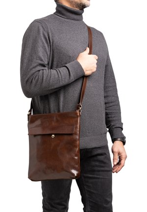 Borsa a tracolla in pelle marrone con una superficie liscia, tasca frontale piatta e accessori dorati. Indossata sopra un maglione grigio e pantaloni neri.