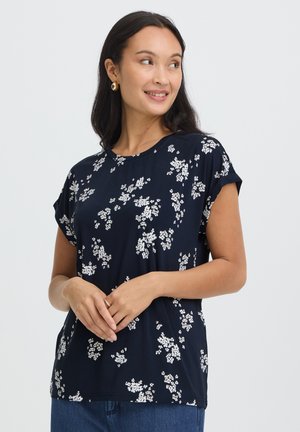 Femme aux longs cheveux foncés portant des boucles d'oreilles en or et un haut bleu marine à manches courtes avec de petits motifs floraux blancs, debout devant un fond uni.