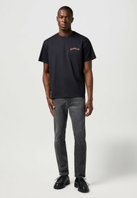 Camiseta de algodón negra con el logo rojo "WRANGLER?" en el lado izquierdo del pecho, combinada con jeans de mezclilla grises y zapatos negros con cordones.