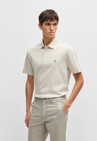Lys beige kortermet polo med krage, tre knapper og en liten logo på brystet, sammen med lysegrå bukser.