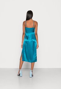 Vestido midi de cetim em tom de verde-água, com alças finas, fenda nas costas, textura suave e uma silhueta solta. Combinado com sandálias de salto bloco em azul claro.