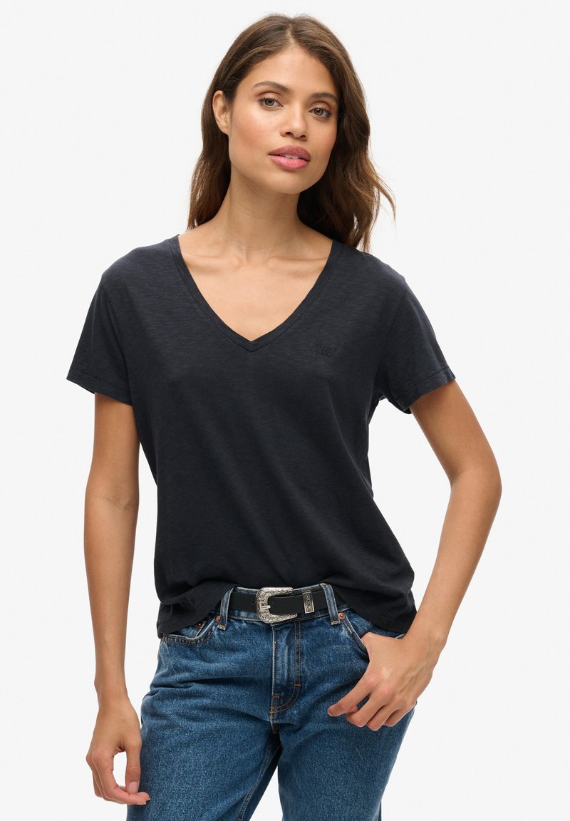 Camiseta de cuello en V de un azul marino oscuro, fabricada en un material ligero, con mangas cortas y un logo sutil en el pecho, combinada con unos vaqueros azules y un cinturón.