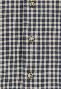Overhemd met knopen, uitgevoerd in een marineblauw en crèmekleurig ruitpatroon. Gemaakt van geweven katoen, met ronde, tankleurige knopen langs de knoopsluiting.
