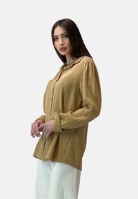 Camicia a maniche lunghe con bottoni in tessuto texturizzato color marrone chiaro, con colletto morbido e vestibilità ampia, abbinata a pantaloni bianchi.