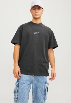 Sort oversized t-shirt med lilla "PREMIUM STUDIOS" tekst i midten, parret med lyseblå cargo jeans. Stoffet ser blødt ud.