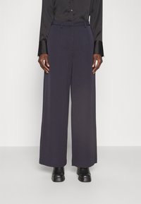 Pantalon à jambes larges en tissu bleu marine foncé, doté d'une taille haute, de passants pour ceinture et d'une texture lisse. Porté avec des bottines noires.