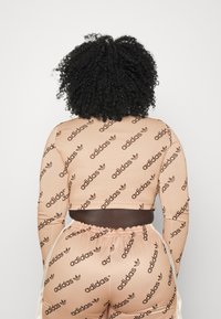 Osoba s kudrnatými vlasy viděná zezadu, oblečená v béžovém setu Adidas, který má opakující se černý vzor loga Adidas.