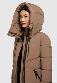 Chaqueta acolchada marrón con una gran capucha, que presenta cremalleras negras, cordones ajustables y bolsillos frontales en diagonal. Textura suave y acolchada.