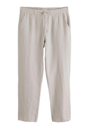 Person trägt hellbeige Leinenhose mit Kordelzug und einen dunkelgrünen, gerippten Pullover, die Hand ruht in der Nähe der Tasche.