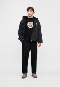 Μαύρο μπουφάν με επένδυση από φλις, μαύρο μπλουζάκι με γραφικό σχέδιο, μαύρο παντελόνι και μπεζ σουέντ παπούτσια. Απλός, καθημερινός σχεδιασμός με έντονη υφή.