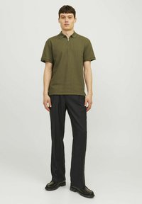 Olivgrön polo-shirt med dragkedja, korta ärmar och en subtil textur, kombinerad med svarta byxor och svarta skor.