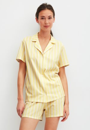 Vrouw die een geel-wit verticaal gestreept kort mouw overhemd met knopen en bijpassende korte broek draagt, staand tegen een effen witte achtergrond.