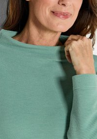 Lichtgroene, geribbelde top met een hoge hals, vervaardigd van een textuurstof en met lange mouwen. Handen die de kraag vasthouden, waarbij de details worden getoond.