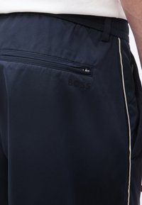 Gros plan sur un pantalon marine avec une poche arrière zippée, un liseré blanc sur le côté et un logo "BOSS" discret sous la poche.