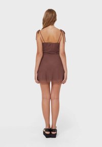 Robe sans manches marron avec des bretelles ajustables, corsage cintré et jupe évasée. Texture lisse, longueur mi-cuisse, portée avec des chaussures à plateforme.