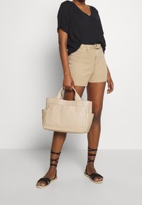 Woman wearing black V-neck top, beige belted shorts, black lace-up sandals, holding a beige leather handbag by one side.