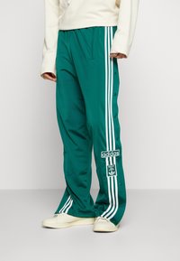 Osoba oblečená v zelených teplákových kalhotách Adidas s bílými bočními pruhy a cvoky, spárovaná s bílými teniskami a béžovým topem s dlouhým rukávem.