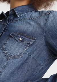 Denimskjorta med klassisk krage, knappstängning och bröstficka med silverknäpp. Mörkblå färg med en texturerad, sliten yta.