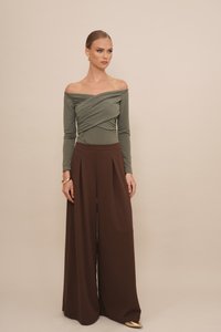 Oliwkowa bluzka typu off-shoulder z drapowaną linią, zestawiona z szerokimi, wysokimi spodniami w kolorze brązowym. Złota biżuteria podkreśla dłonie.