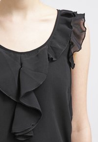 Haut noir avec accents volantés autour du col et des épaules. Confectionné dans un tissu doux à la texture lisse et à la coupe décontractée.