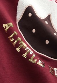 Maroon sweatshirts met een bruine chocoladecake-ontwerp met witte stippen, gouden pailletten tekst met de tekst "EEN BEETJE." Gestructureerde stofaccenten.