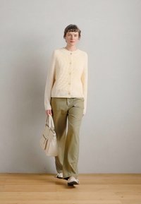 Κρεμ καπιτονέ κάρντιγκ με κουμπιά, ελιά πράσινα παντελόνια και μπεζ τσάντα. Το μοντέλο φοράει ανοιχτά παπούτσια, με απλό χτένισμα, σε ουδέτερο φόντο.