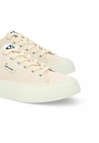 Zapatillas de lona en beige claro con suela y cordones de goma blanca. Presentan un pequeño logotipo en blanco y negro y un diseño de punta redonda.
