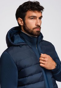 Giubbotto imbottito blu navy con cappuccio, caratterizzato da sezioni trapuntate e tessuto morbido. Chiusura con zip e dettagli a coste sul colletto. Mani posizionate sulla zip.