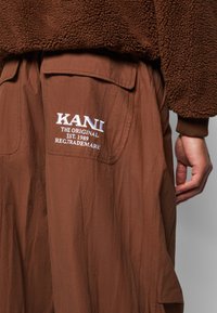 Brune cargo-bukser har en dekorativ lomme med hvid broderet tekst "KANI THE ORIGINAL EST. 1989 REG.TRADEMARK" på bagsiden.