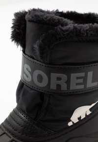 Bota de invierno negra con un cuello de pelaje sintético negro y esponjoso, parte superior de tela, suela de goma y una correa gris prominente que lleva el nombre de la marca "SOREL".