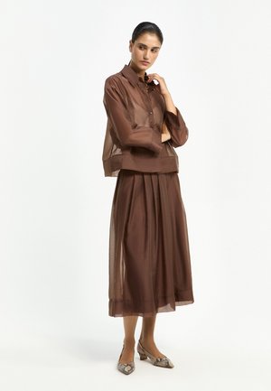 Woman wearing a sheer brown long-sleeve blouse and matching midi skirt with snakeskin-patterned low heel shoes, standing with arms crossed.