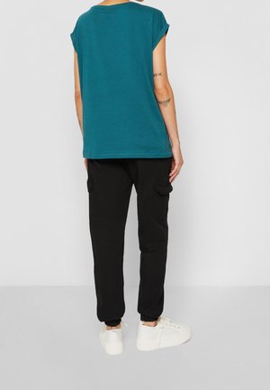 Persoon die een tealkleurig T-shirt met korte mouwen, zwarte cargobroek met zakken en witte sneakers draagt, staand met de rug naar de kijker tegen een effen achtergrond.