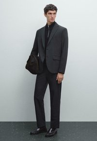 Homme portant un costume noir sur mesure, un pull zippé noir, des mocassins noirs, tenant un sac en suedine marron foncé, se tenant contre un mur gris clair.