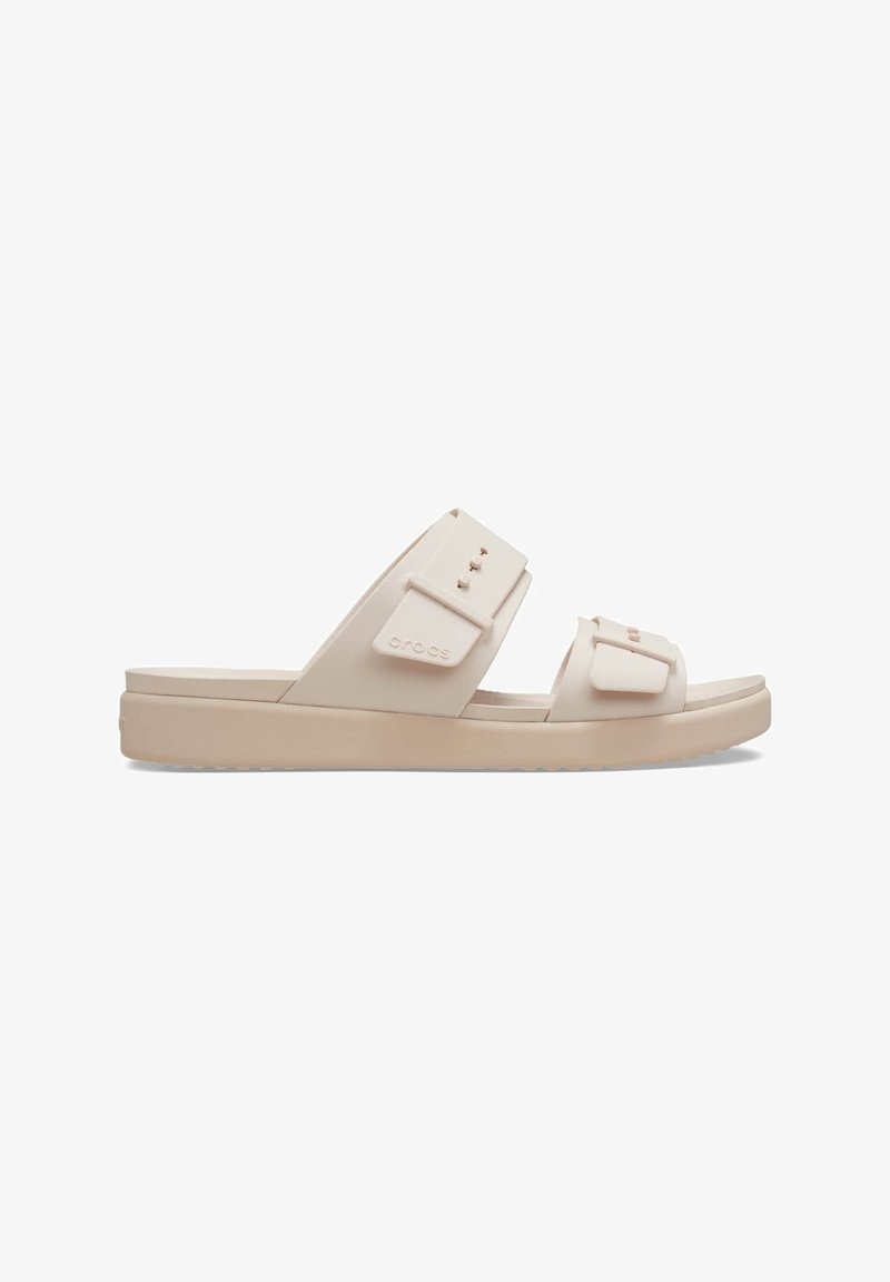 Beige slide sandals with two adjustable straps, flat sole, and textured tread. Made from soft material, featuring a logo and decorative accents.