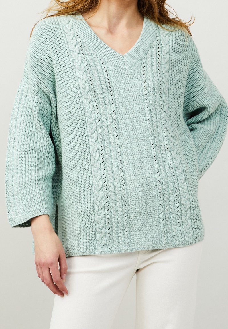 Femme portant un pull ample bleu clair à col en V en maille torsadée avec des manches trois-quarts et un pantalon blanc, debout devant un fond uni.