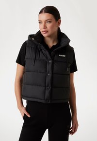 Gilet imbottito nero con cappuccio, caratterizzato da un design a quadri, bottoni frontali e un logo. Indossato sopra una maglietta nera, abbinato a pantaloni neri.