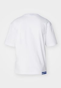 Camiseta blanca de algodón con cuello redondo, mangas cortas y una etiqueta de marca azul en la parte inferior izquierda. Textura suave.