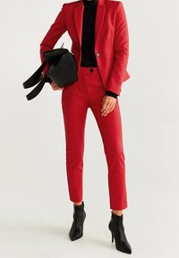 Blazer rouge et pantalon assorti en tissu lisse, associés à un col roulé noir et des bottines noires, tenant un sac noir.