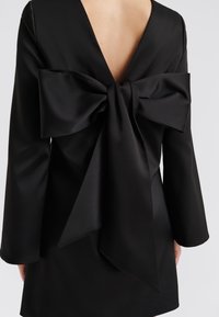 Svart satengkjole med dyp V-rygg, utstyrt med en stor sløyfe i midjen og lange ermer. Glatt tekstur med en strukturert design.