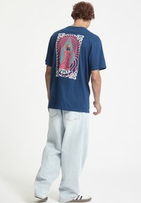 Kongelig blå bomulds t-shirt med et grafisk design, der fremviser abstrakte linjer og en figur på ryggen. Løs pasform, parret med lyse denimbukser.