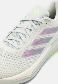 Gros plan d'une chaussure de course en mesh gris clair avec des rayures lavande, des lacets blancs et un logo Adidas vert néon sur la languette.