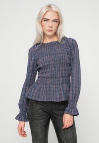 Fiatal nő hosszú szőke hajjal, sötétkék és piros kockás, smokkos blúzt és fekete textúrált nadrágot visel, egy egyszínű háttér előtt áll.