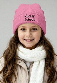 Pink knit beanie with a rectangular patch labeled "Zucker Schock." Paired with a white, textured scarf and a light beige jacket.